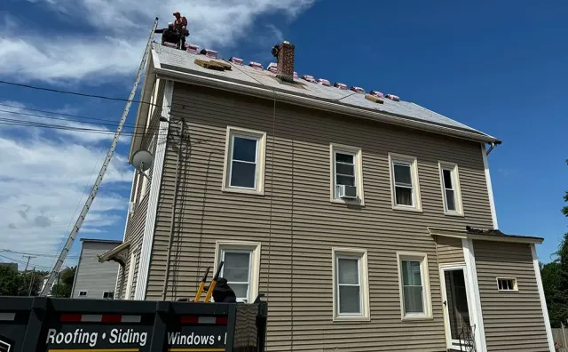 Roofers on a house replacing the roof on a sunny day.  Ladder, dumpster, and blue sky visible.