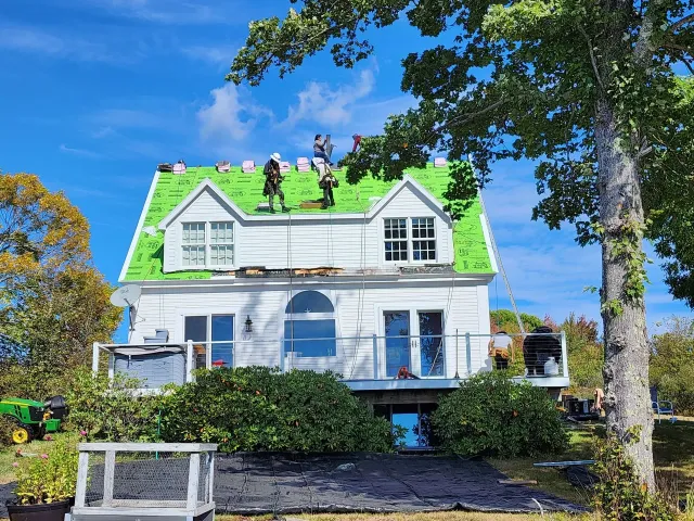 White house with green roof under construction; workers on roof in sunlight.