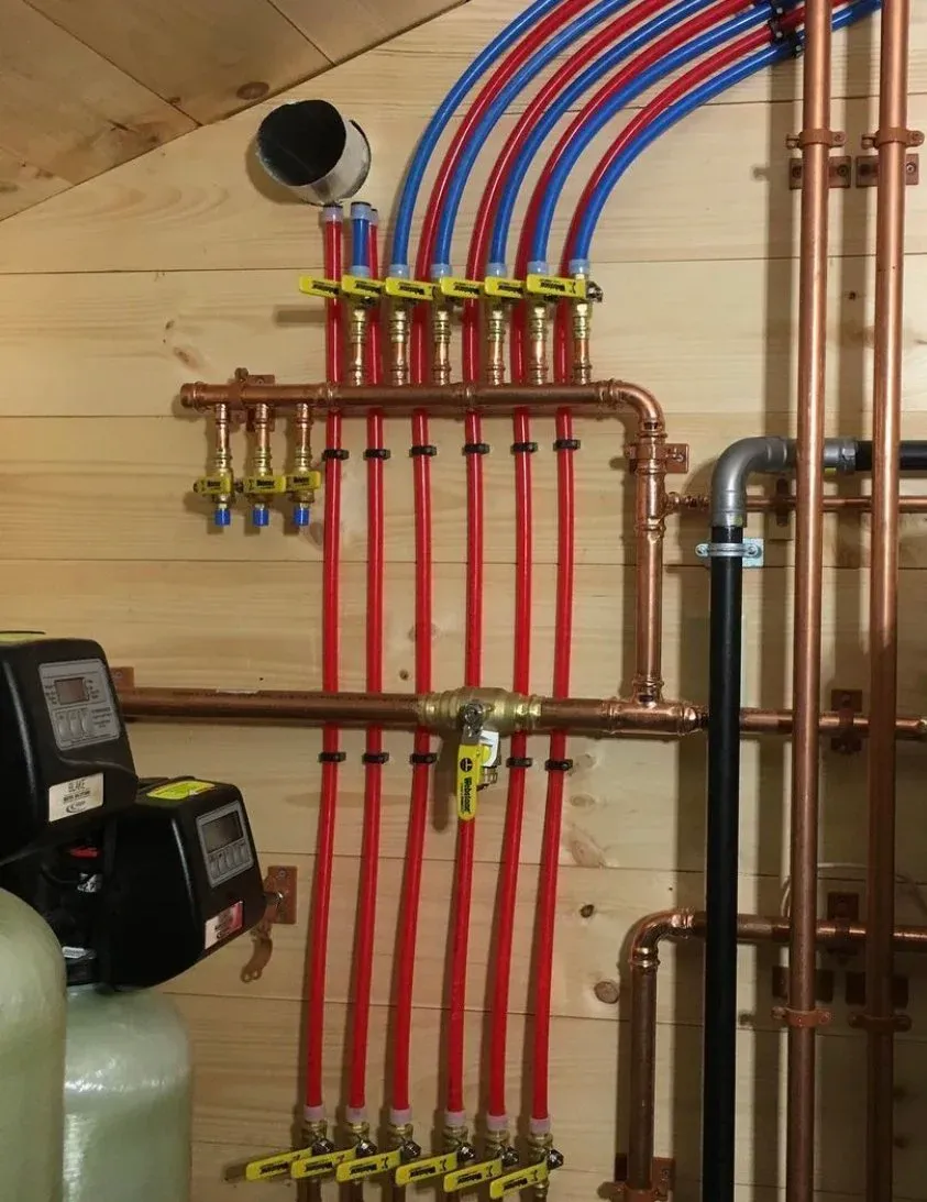 Plumbing manifold with red and blue PEX pipes in a wooden-walled room, featuring yellow shut-off valves, copper pipes, and water treatment equipment.