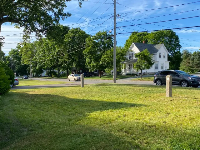 Residential street with cars, grass, and large trees under a blue sky.