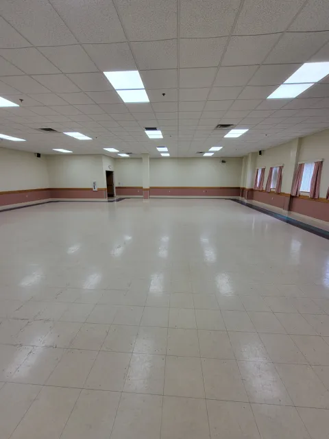Empty banquet hall or event room with tiled floor, beige walls, pink and wooden trim, ceiling tiles with fluorescent lights, and windows with pink curtains.
