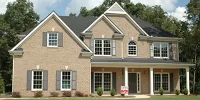 A two-story tan brick house with a dark shingled roof, grey shutters, a front porch, and a small yard with green trees.