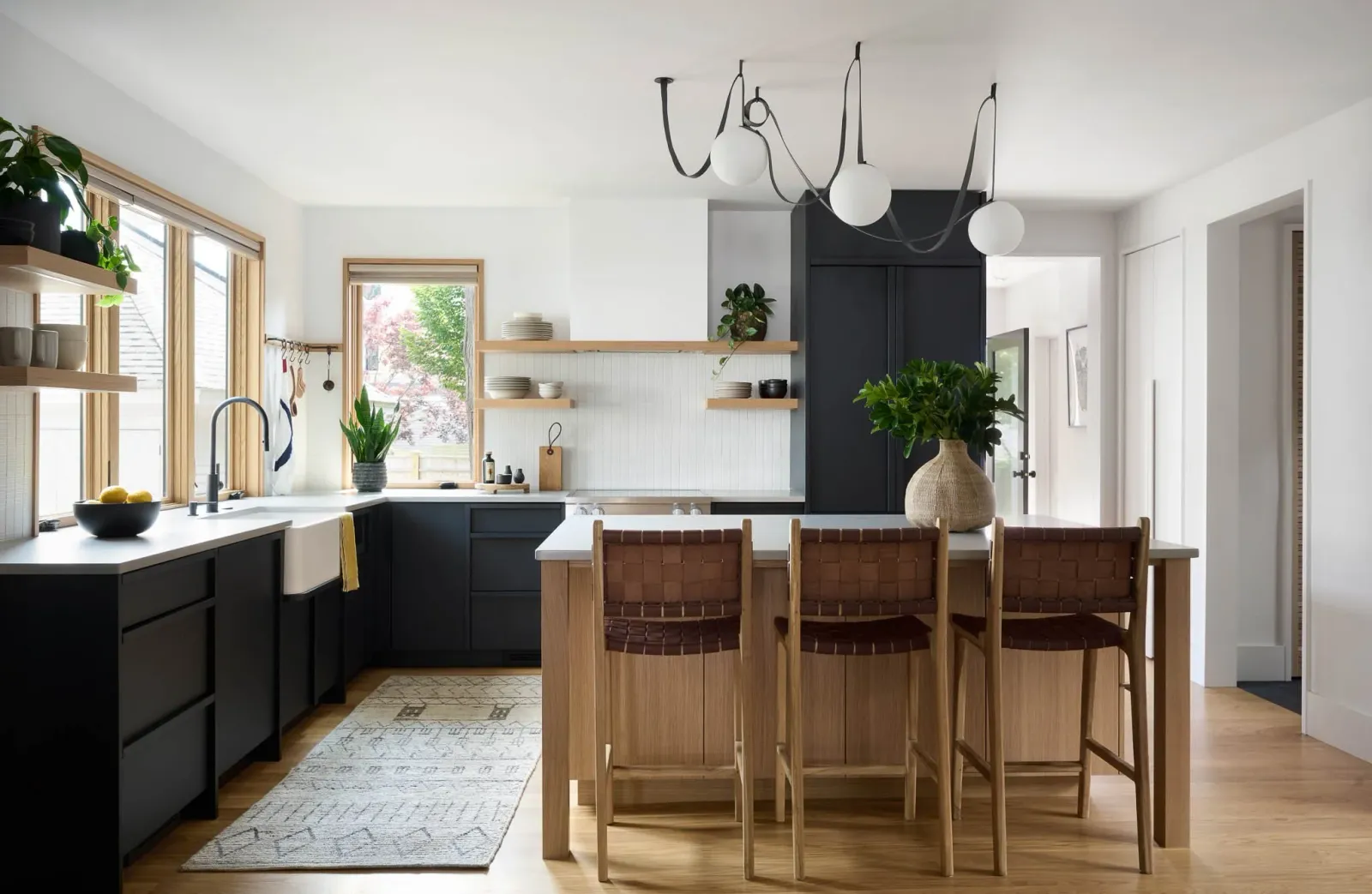 Modern kitchen with black cabinets, wooden accents, and open shelving, featuring a kitchen island with four wooden chairs, a window with plants, and a contemporary light fixture.