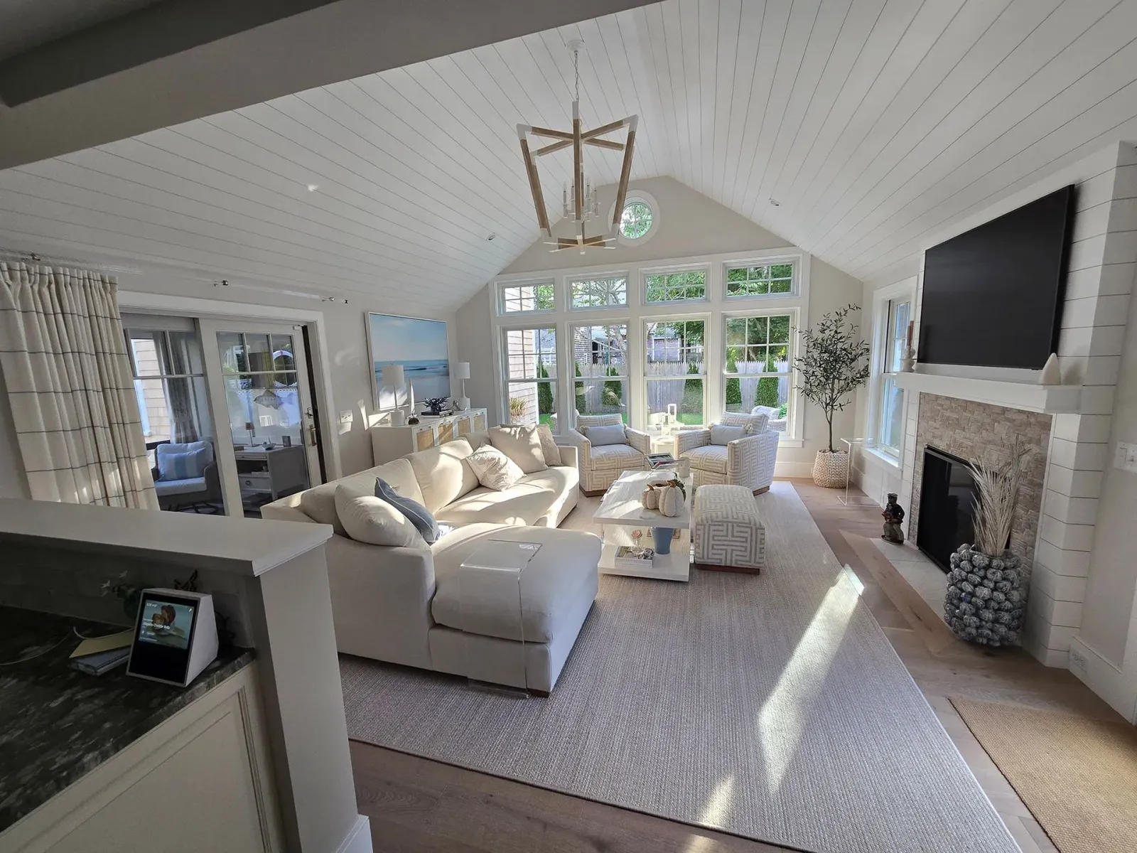Bright living room with white sofas, stone fireplace, large windows, and a TV on the wall.