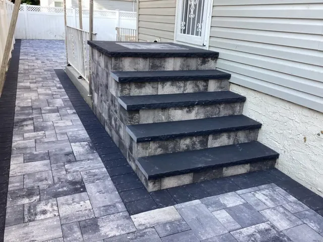 Outdoor side yard with dark steps, black railing, and stone paver walkway beside a house