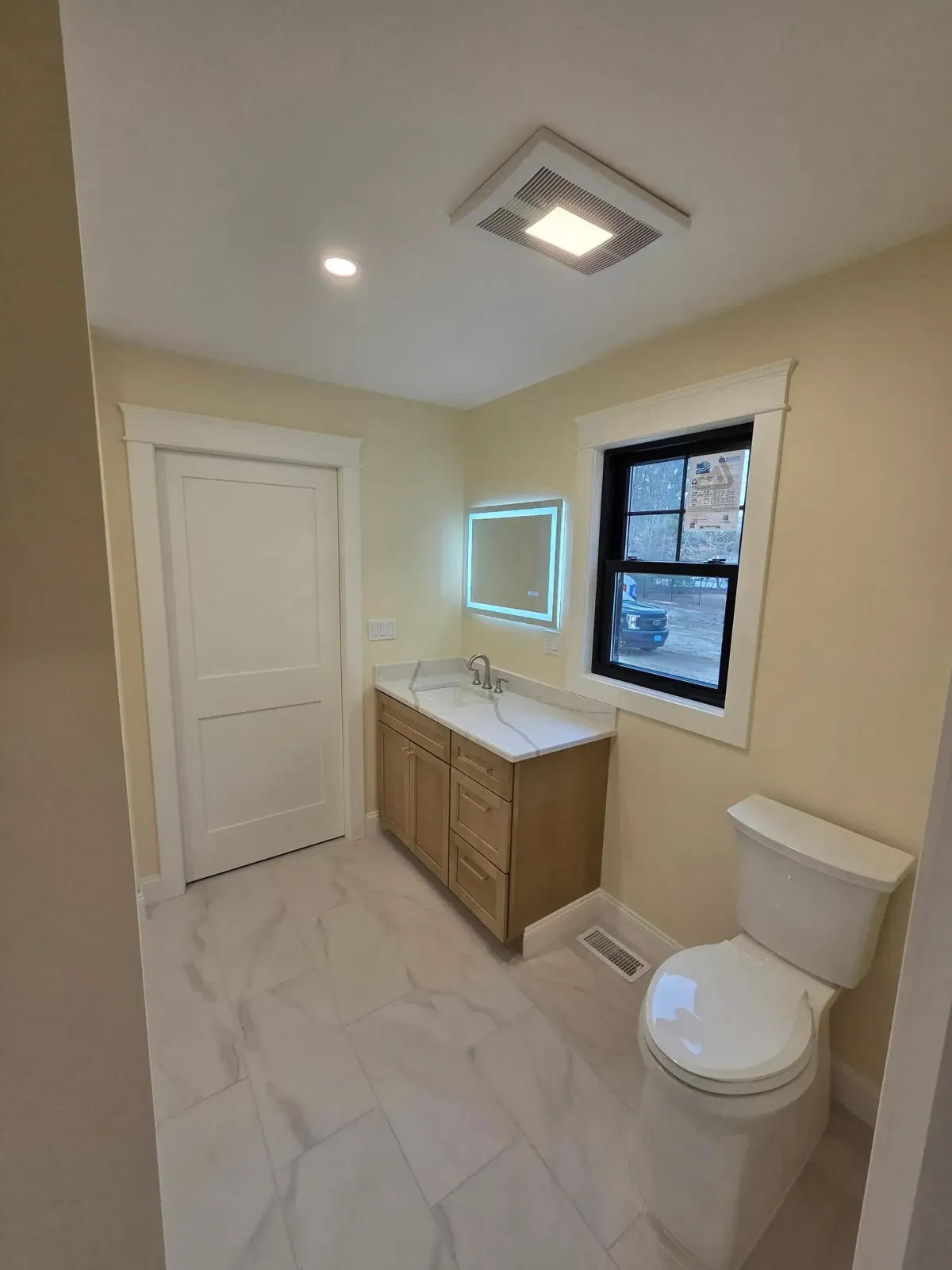 Bathroom with light beige walls, marble tile floor, wood vanity, white toilet, and window with black trim.