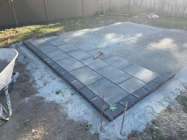 A partial patio installation with dark stone pavers bordered by a darker row of bricks, set on a gravel base in a yard.