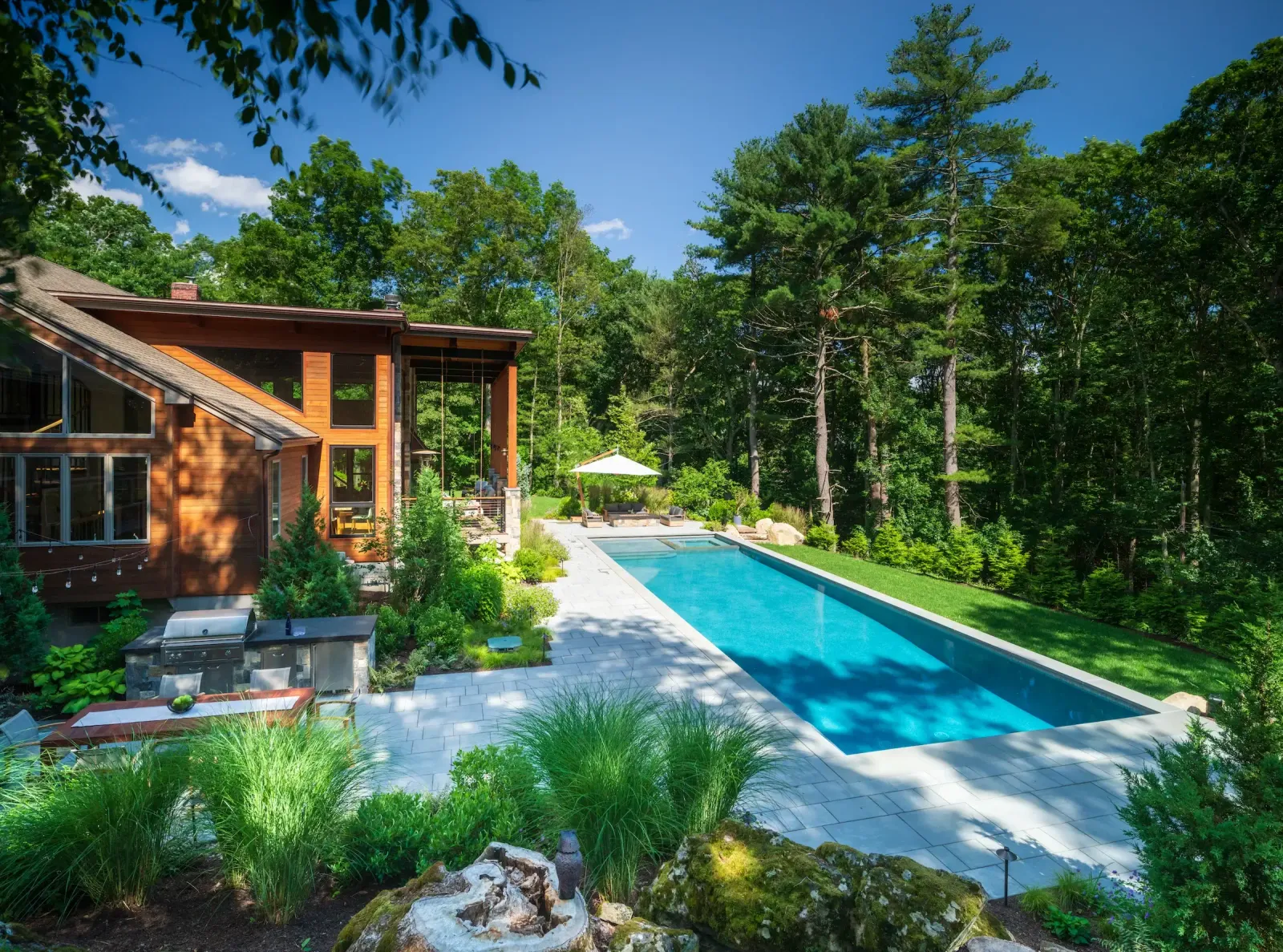 A wide-angle shot of a Wayland, MA backyard featuring a long, rectangular lap pool with light blue water. To the left, a modern wood-sided home overlooks an outdoor kitchen and dining area. The landscape is framed by ornamental grasses and a dense forest of tall pine trees.