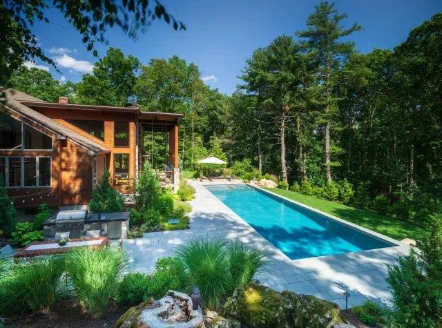 A wide-angle shot of a Wayland, MA backyard featuring a long, rectangular lap pool with light blue water. To the left, a modern wood-sided home overlooks an outdoor kitchen and dining area. The landscape is framed by ornamental grasses and a dense forest of tall pine trees.