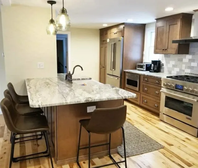 Modern kitchen with a marble island, wooden cabinets, stainless steel refrigerator and stove, small appliances, pendant lights, hardwood floor, and a rug under the island.