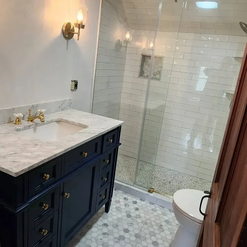 Modern bathroom with a marble countertop vanity, gold fixtures, wall sconce, glass-enclosed shower with white subway tiles, and a toilet.