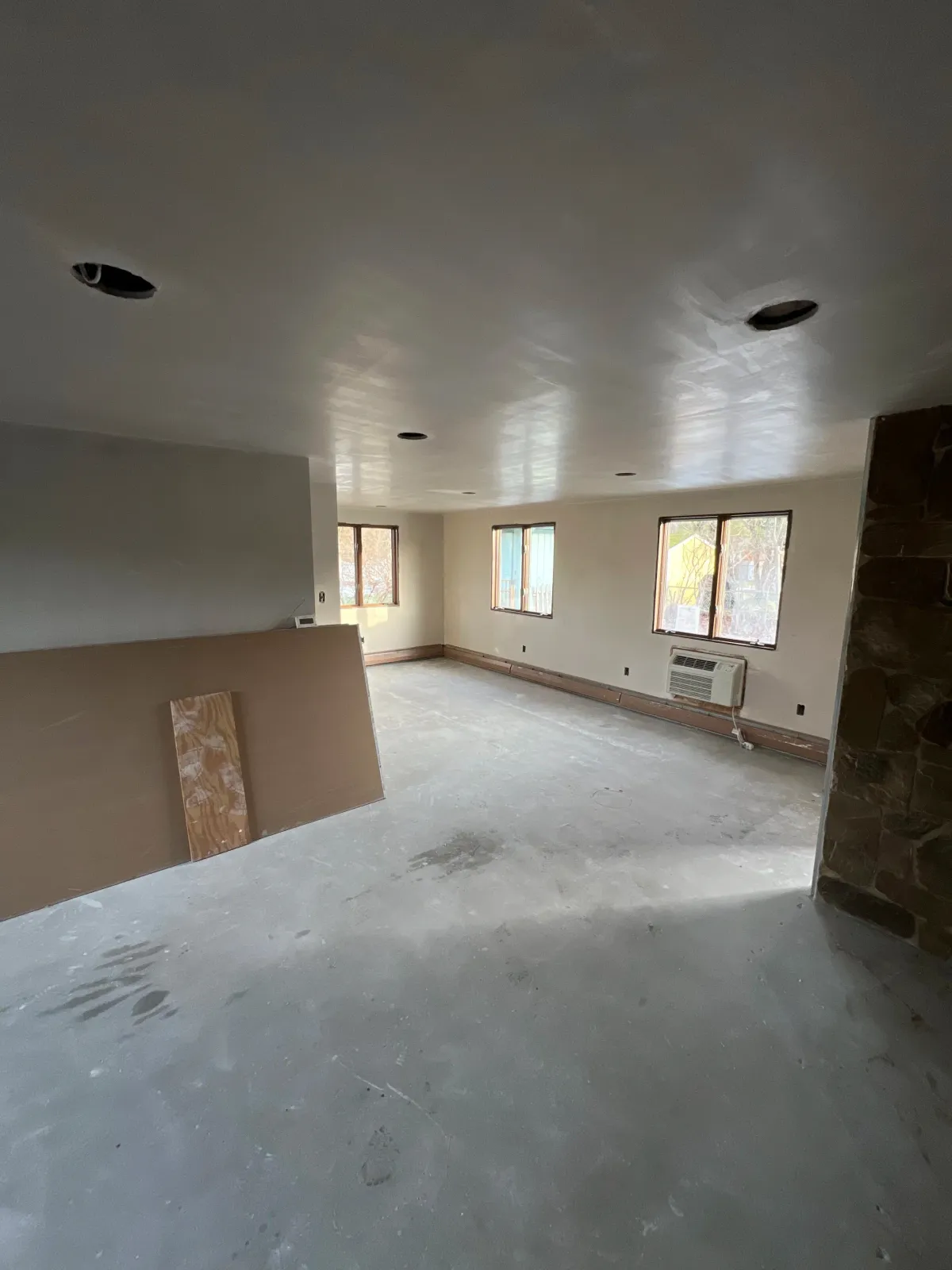 Empty living room under renovation with unfinished floor, four windows, and a wall-mounted air conditioning unit.