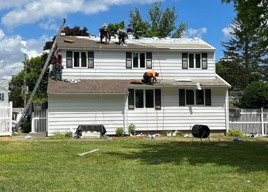 Roofing crew working on a two-story white house with black shutters on a sunny day.