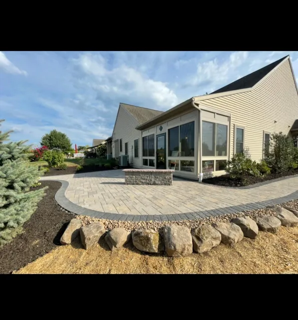 Backyard patio area with stone pavers, landscaped plants, and a screened porch attached to the beige house under a blue sky with clouds.