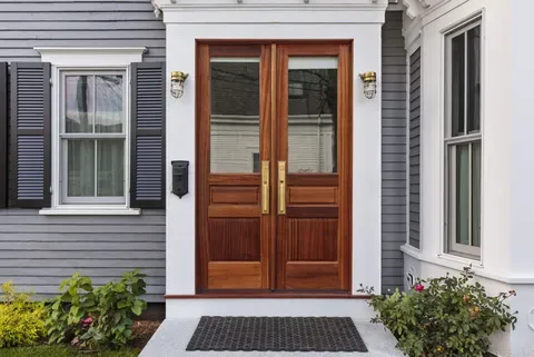 Front entrance with wooden double doors, white trim, gray siding, and potted plants by the steps.