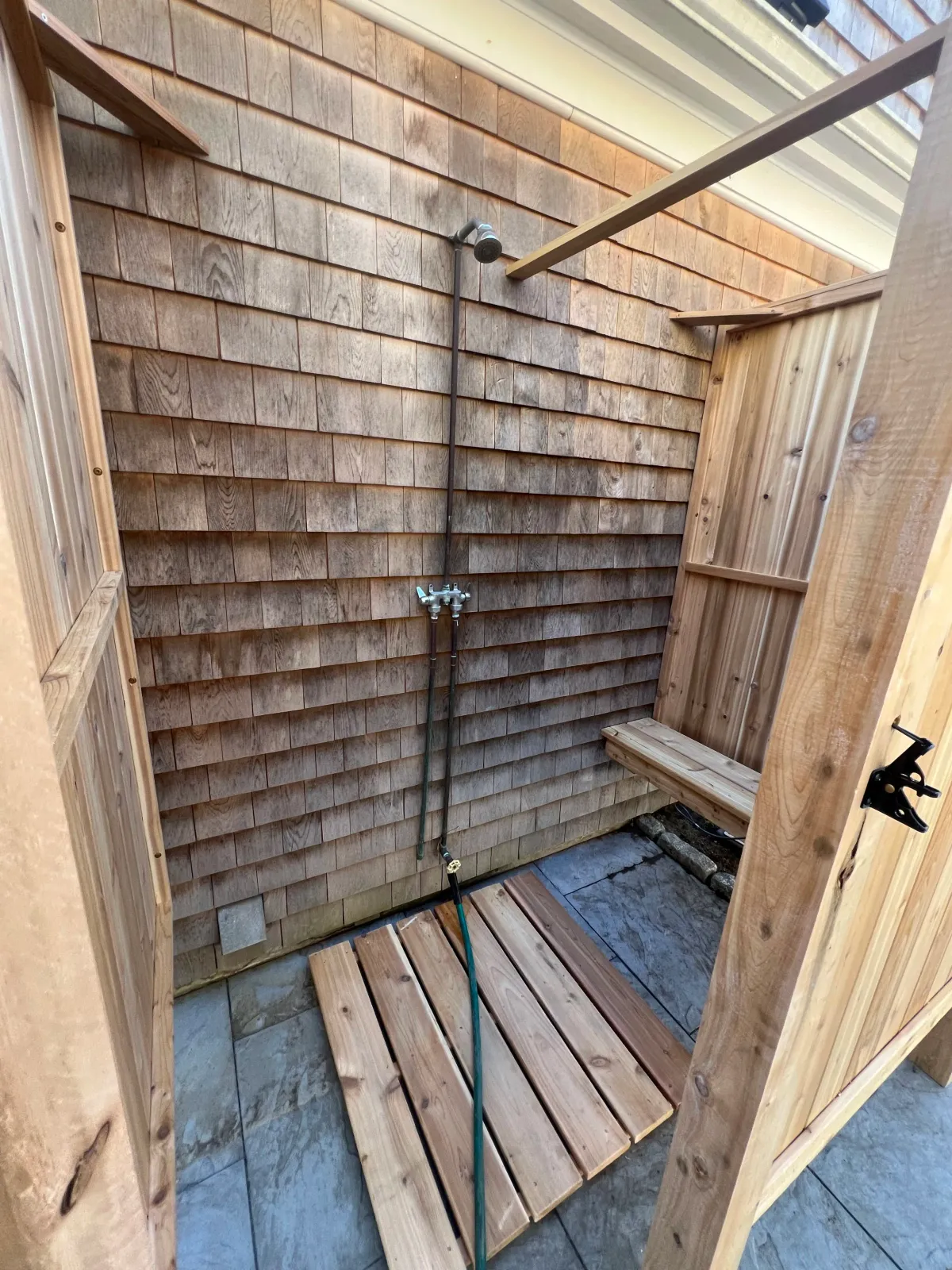An outdoor shower area under construction with wooden walls, a showerhead, a hose, a small wooden bench, and a wooden platform on a tiled floor.