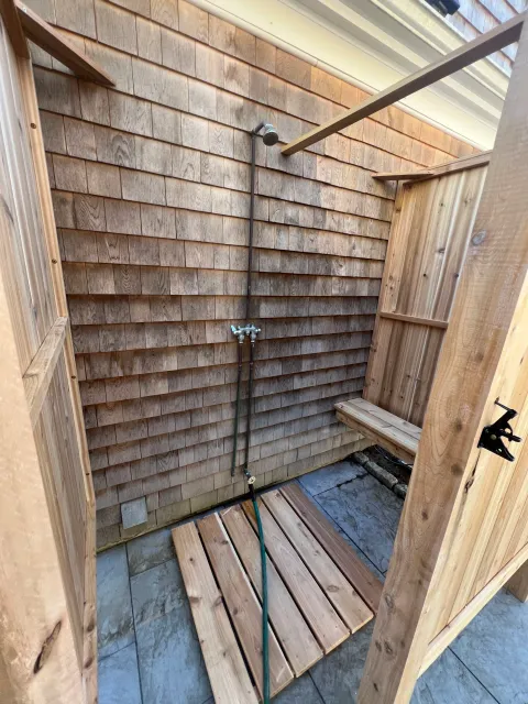 An outdoor shower area under construction with wooden walls, a showerhead, a hose, a small wooden bench, and a wooden platform on a tiled floor.