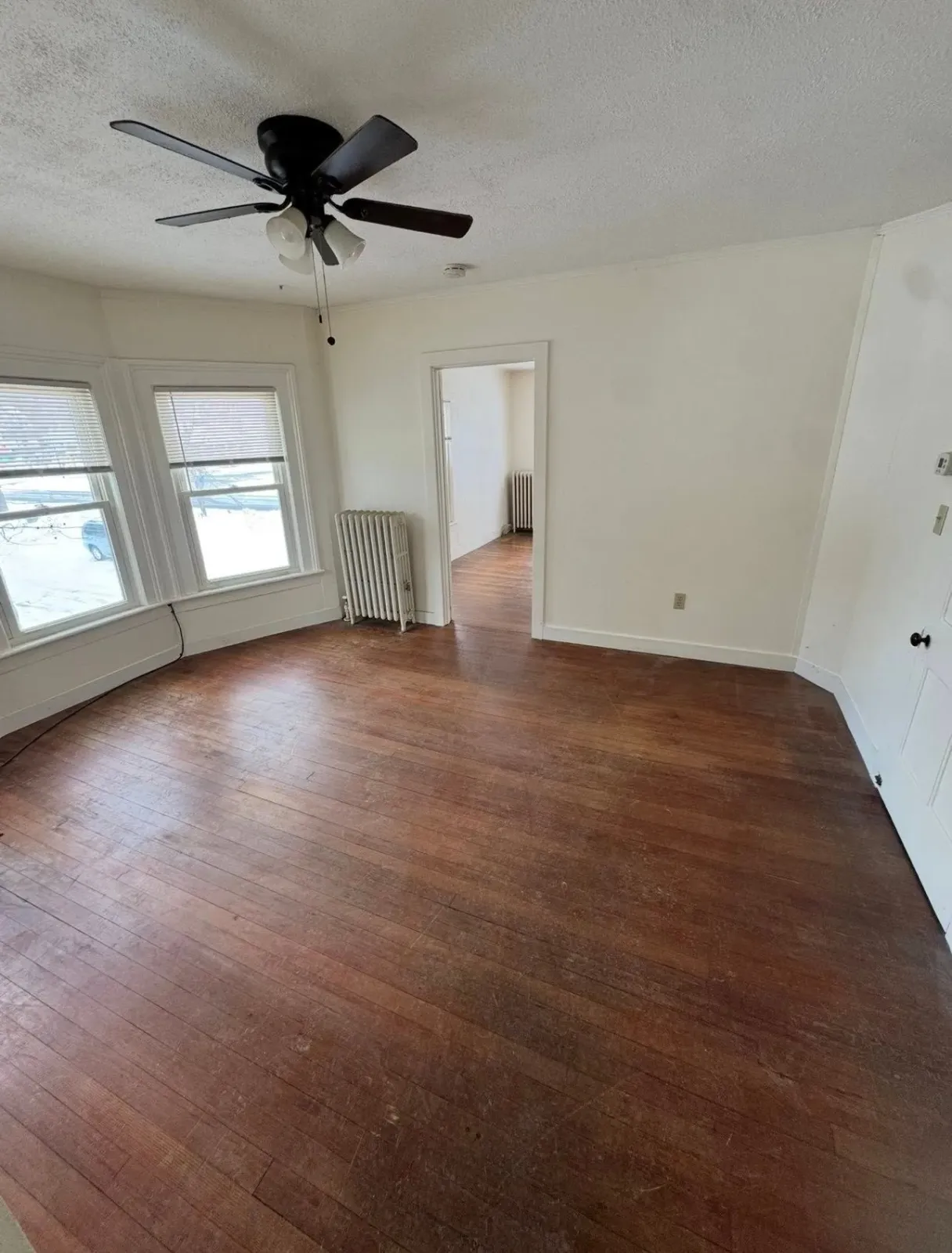 An empty room with dark wood flooring, beige walls, two windows with blinds, a ceiling fan, and a radiator.