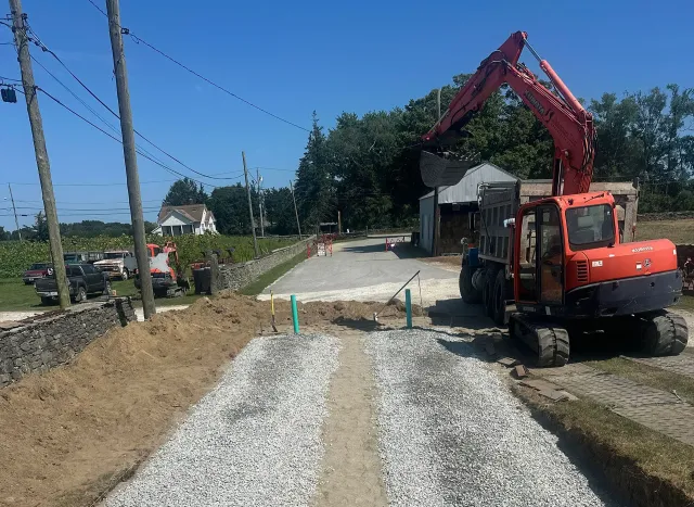 A red excavator stands on a partially graveled driveway at a construction site with trees and utility poles in the distance.