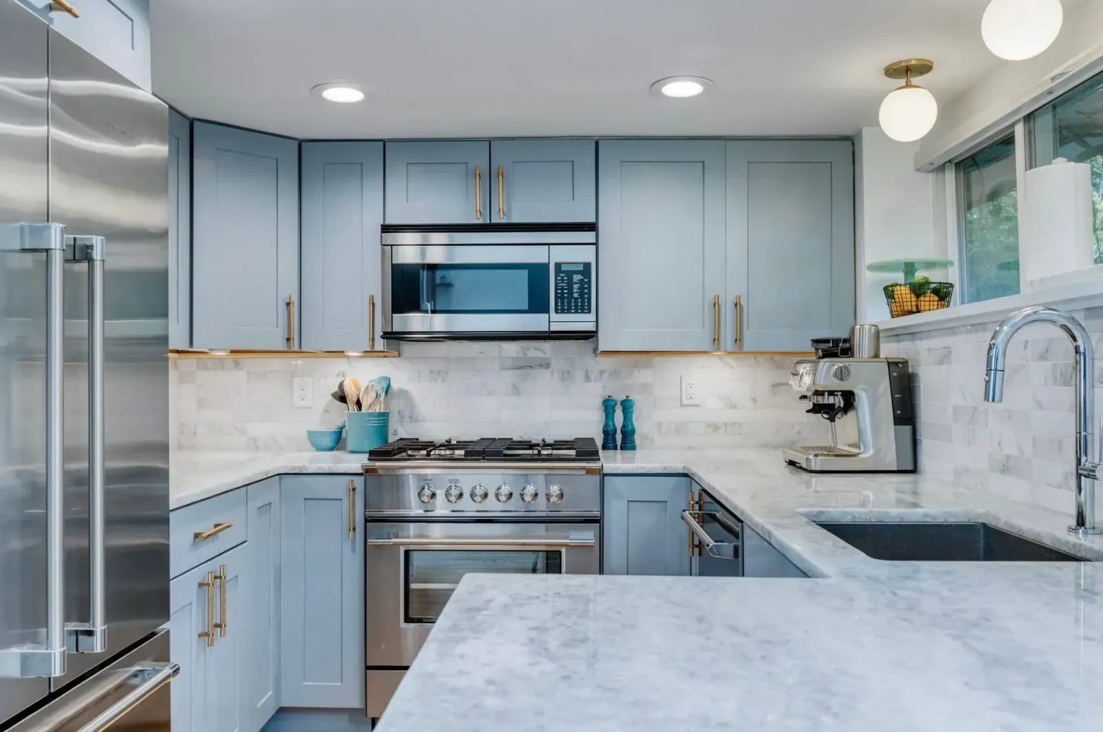 Bright modern kitchen with light blue cabinets, stainless appliances, marble counters, and large windows.