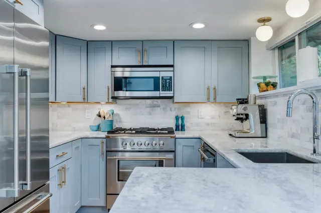 Bright modern kitchen with light blue cabinets, stainless appliances, marble counters, and large windows.