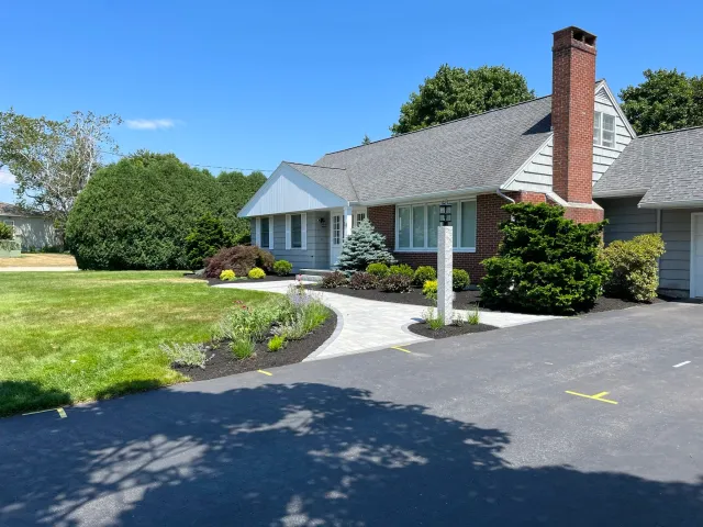 A residential house with a brick chimney, grey siding, and a well-maintained yard featuring shrubs and colorful flowers, along with a paved walkway and parking area.