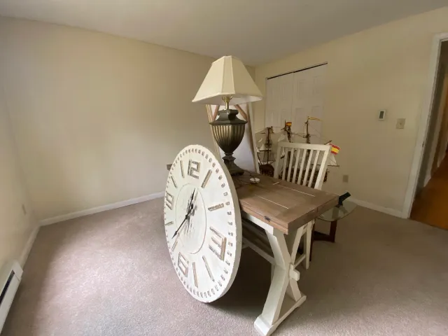 Empty room with a wooden table, a large wall clock leaning against the table, a table lamp, and a model ship in the background.