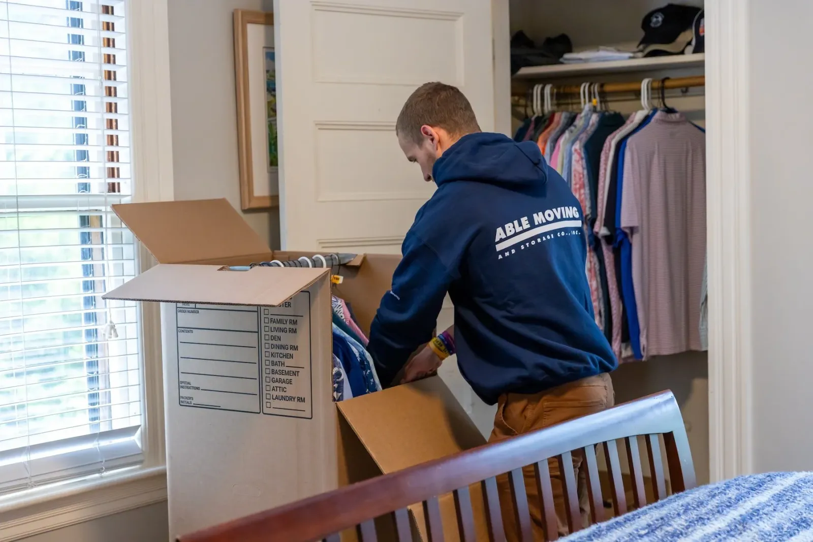 A man packing clothes into a box in a bedroom closet.