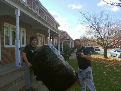 Two men are carrying a large black object in front of a brick building.