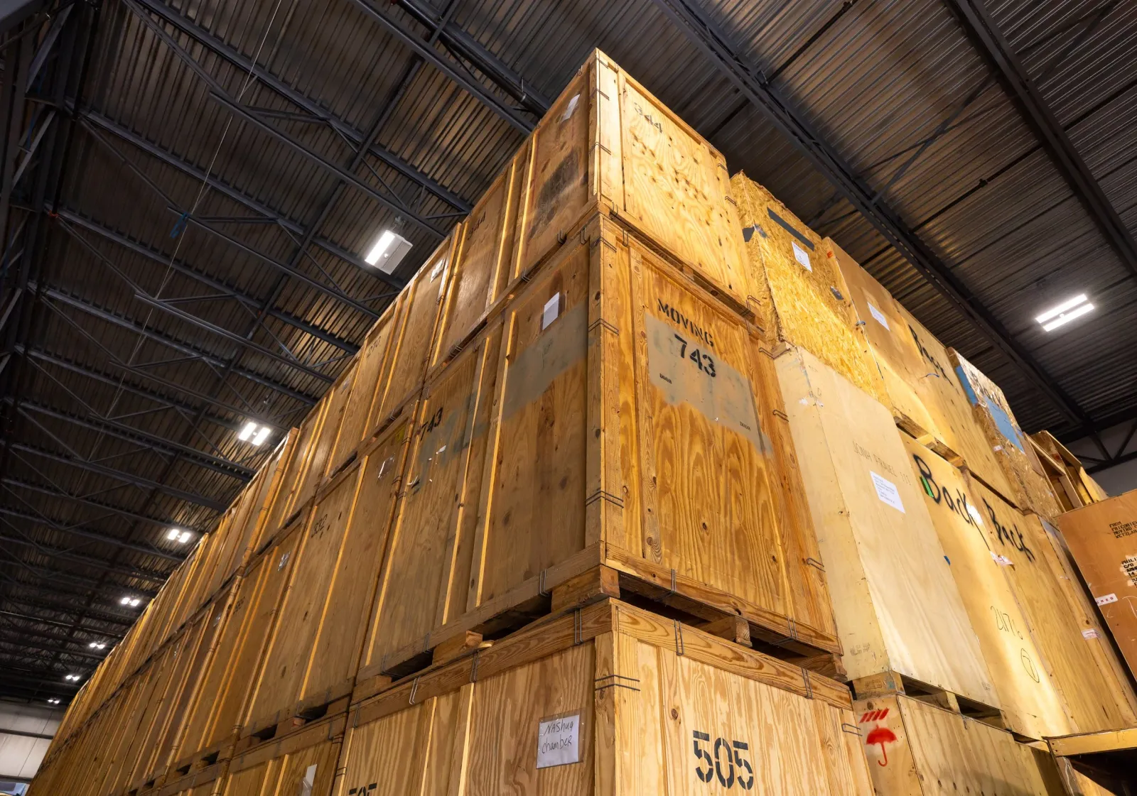 Large wooden crates stacked inside a warehouse with a metal ceiling and bright overhead lights.
