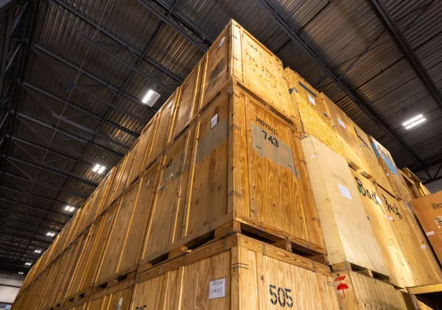 Large wooden crates stacked inside a warehouse with a metal ceiling and bright overhead lights.