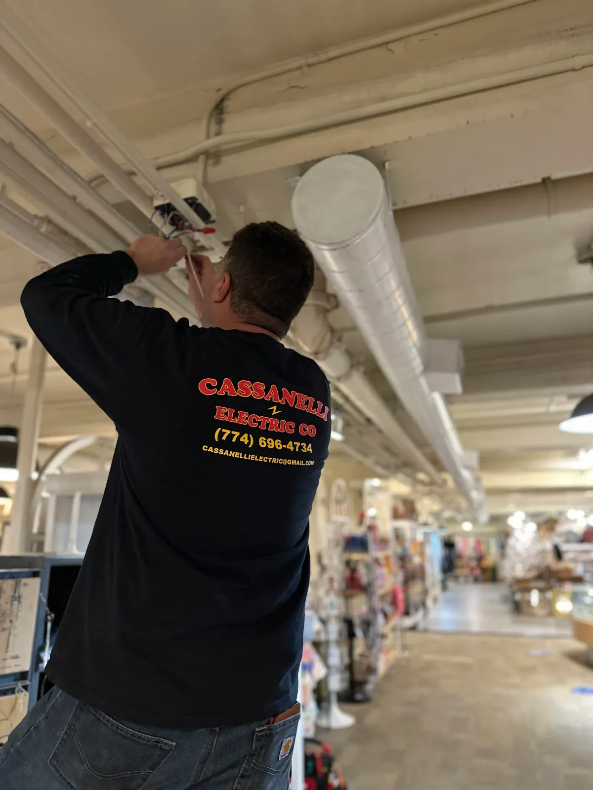 A man working on electrical wiring on the ceiling of a store or warehouse. He is wearing a black shirt with red and yellow lettering on the back that reads 'Cassanelli Electric Co' with contact info. Large industrial ductwork is visible on the ceiling.
