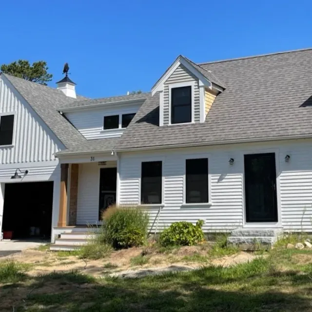 Front view of a white house with a gray shingled roof, two stories, and a small front porch. There are three black-framed windows on the first floor and three similar windows on the second floor, with a dormer window. A small grassy area with bushes is in front of the house. Part of another building with a similar siding is visible on the left. Clear blue sky in the background.