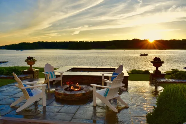 A lakeside setting at sunset featuring two Adirondack chairs beside a lit fire pit, with a boat on the water and trees in the distance.