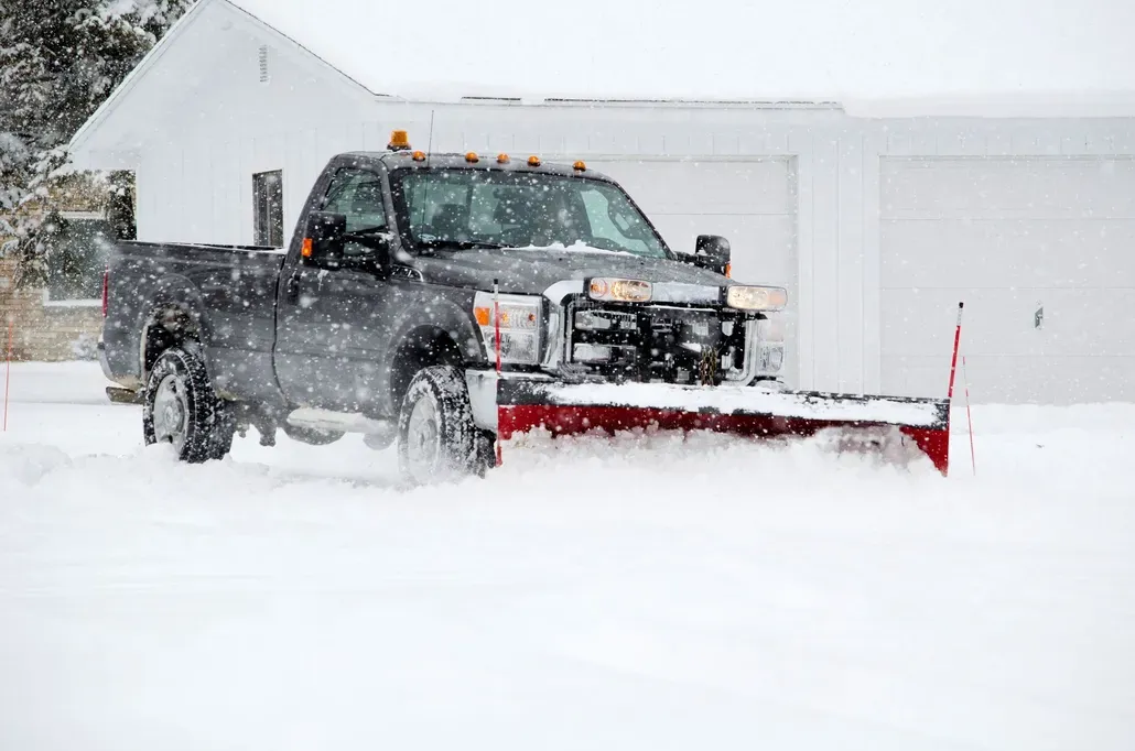 A dark-colored pickup truck with a large, red-bladed snowplow clearing a snow-covered residential driveway.