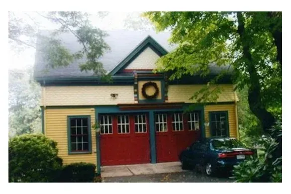 Yellow house with red double doors, dark roof trim, and a parked car in front amid trees