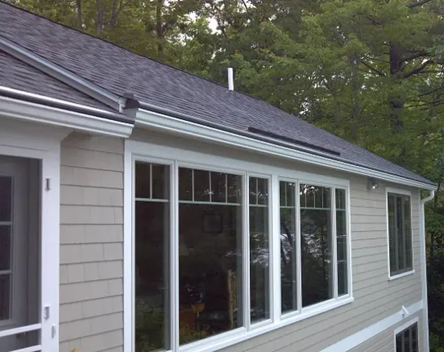A side view of a house with light tan shingle siding, white trim windows, and a dark grey shingled roof.