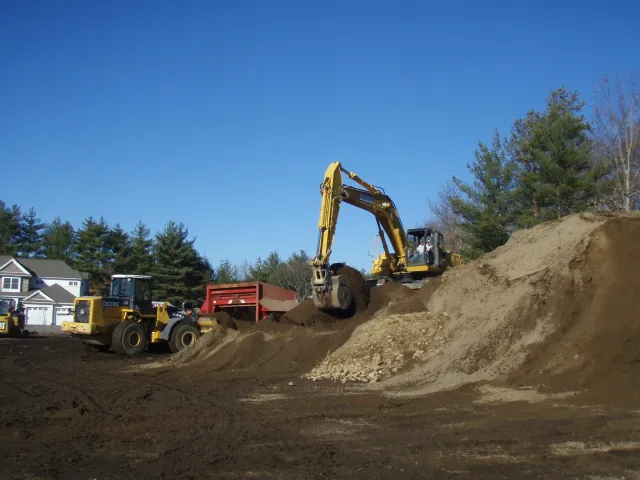 Construction site with a yellow excavator digging into the dirt, residential houses in the background, and trees under a blue sky.