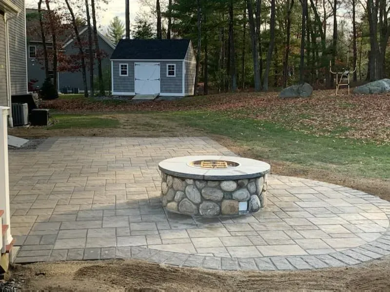New paver patio with a custom stone fire pit surrounded by a stone wall, in a yard with a shed in the background.