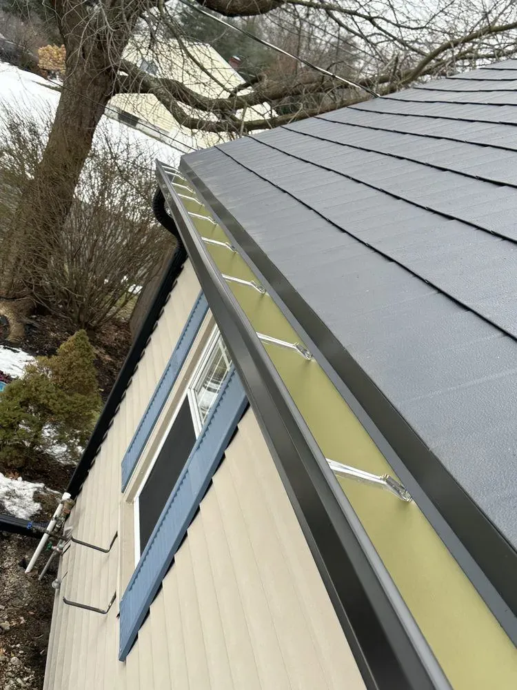 Tilted view of a house roof with gray metal panels, yellow eaves, and leafless trees nearby