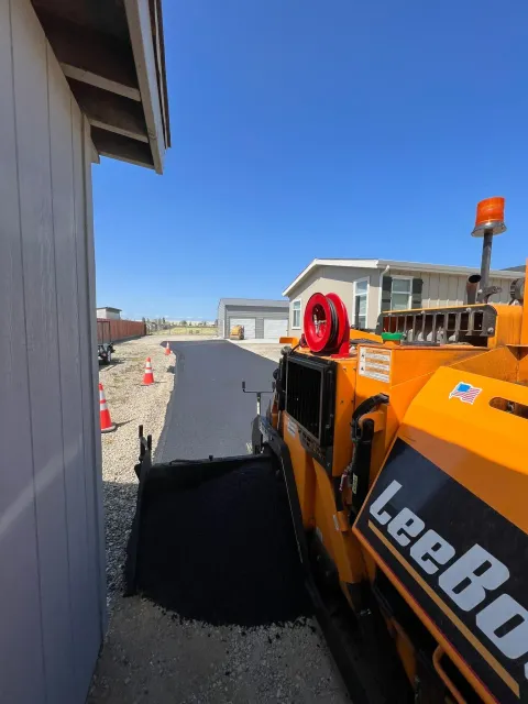 Asphalt paving machine laying asphalt on a driveway next to a building under a blue sky.