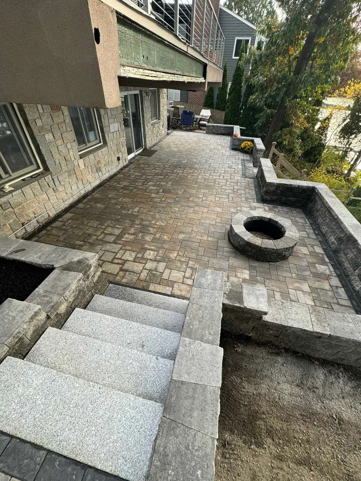 A stone patio with a built-in fire pit and gray stone steps leading up to a house under renovation.