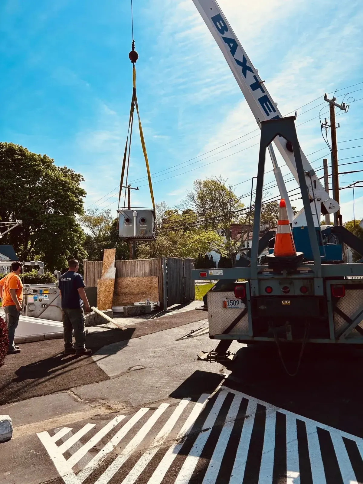 Crane lifting electrical equipment at a construction site on a sunny day. Workers in sight.