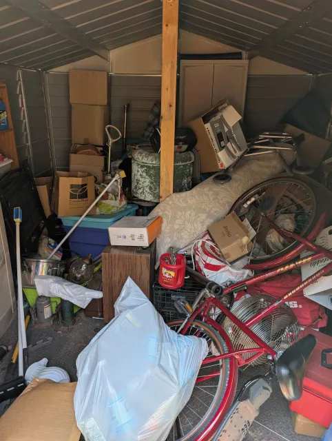 Cluttered storage shed packed with boxes, bags, tools, and bicycles under a slanted roof.
