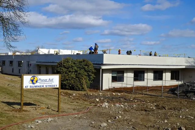 The Planet Aid Headquarters under construction. Workers on the flat roof of the building, blue sky. Sign in front: Planet Aid.