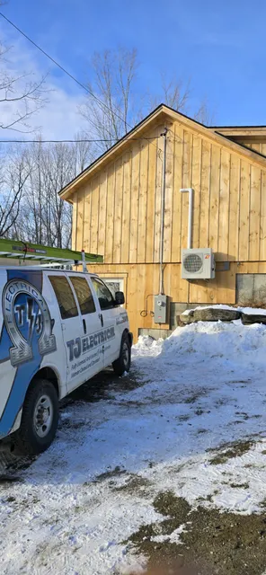 A van with TJ Electrical and Radon Air branding is parked beside a wooden building. Snow covers the ground. An outdoor electrical unit is mounted on the building's wall.
