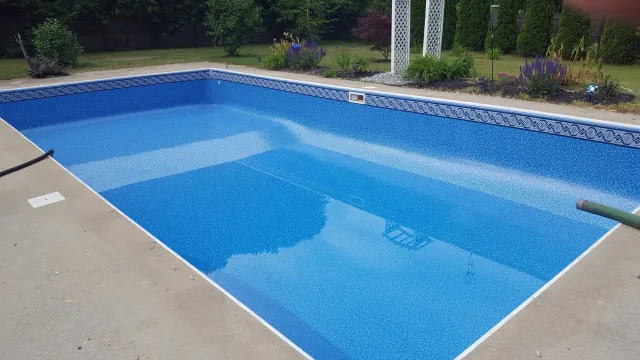 Empty in-ground swimming pool with blue mosaic tiles, surrounded by a concrete deck and landscaped garden with trees, bushes, and purple flowers.