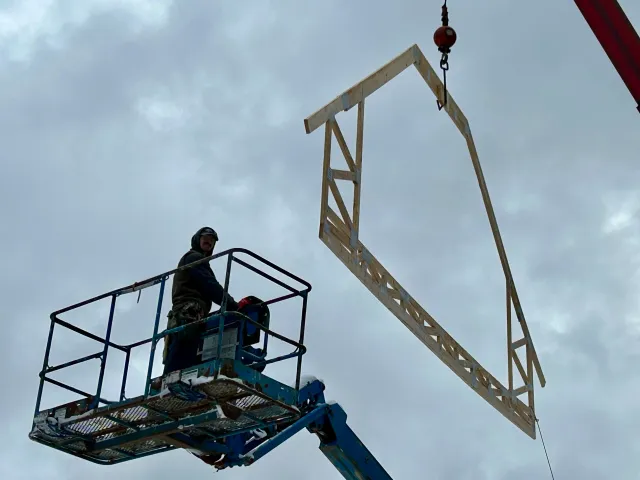 A worker on a lift is installing a framed piece of the a house, suspended by a crane.
