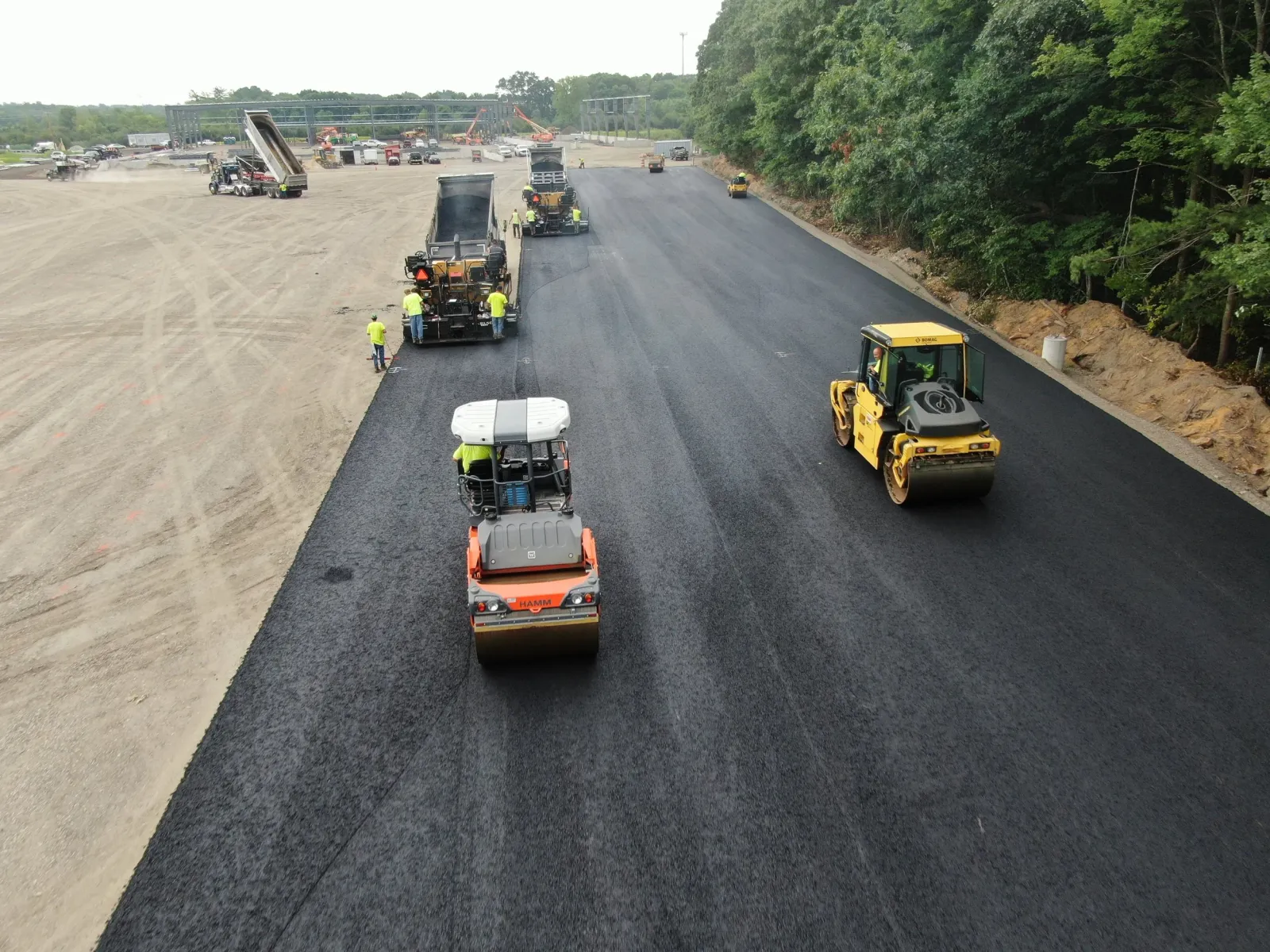 Construction workers are paving a new road with asphalt, using heavy machinery and rollers to flatten and smooth the surface. The scene includes multiple workers wearing bright yellow safety vests and helmets, and there is a partially completed section of road next to a wooded area.