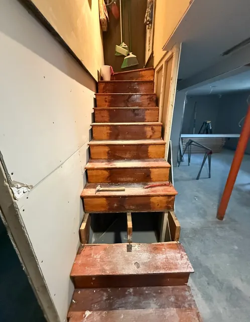 Staircase mid-demolition with exposed treads, risers, and framing during a custom carpentry rebuild